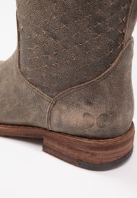 Botte de cowboy en daim marron avec une tige texturée ornée de motifs croisés, un talon en bois empilé, et une semelle en caoutchouc foncé.