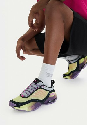 Osoba v podřepu, oblečená v černých šortkách, bílých ponožkách značky Reebok a neonově žlutých a fialových teniskách Reebok, na jednobarevném bílém pozadí.