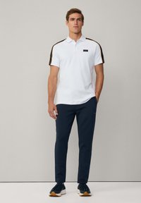 Hvid polo-shirt med sorte og orange striber på skuldrene, parret med navy bukser og sorte sneakers med beige såler.