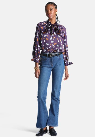 Femme portant un chemisier en satin violet à motif floral avec un nœud papillon, un jean évasé taille haute bleu, une ceinture noire, des mocassins noirs et des bijoux en or.