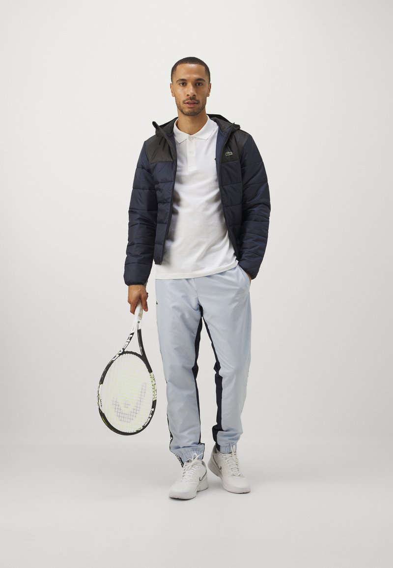 Dicke Jacke in Marineblau mit schwarzen Akzenten, weißes Polohemd, hellblaue Trainingshose mit Seiteneinsätzen, hält ein Tennisschläger, trägt weiße Sneaker.