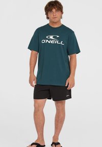 Tamsiai žalia marškinėliai su balta "O'Neill" logo, derinami su juodomis šortais ir juodais sandalais. Laisvas pasiuvimas, trumpomis rankovėmis, pagrindinis dizainas.