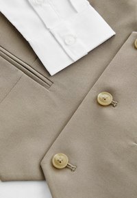 Blazer beige avec deux boutons à l'avant, en tissu texturé, et une chemise blanche à col superposée en dessous, mettant en valeur des lignes épurées et des détails de couture.