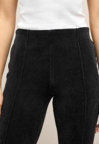 Schwarze Cordhosen mit vertikalem, geripptem Muster, hoher Taille und sichtbaren Nähten, kombiniert mit einem weißen, eng anliegenden Oberteil.