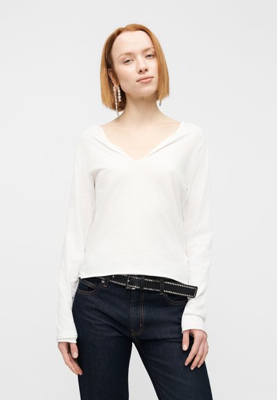 Λευκή μπλούζα μακρυμάνικη με V λαιμόκοψη και ελαφρώς χαλαρή εφαρμογή, συνδυασμένη με σκούρα μπλε τζιν και μαύρη ζώνη με καρφιά.