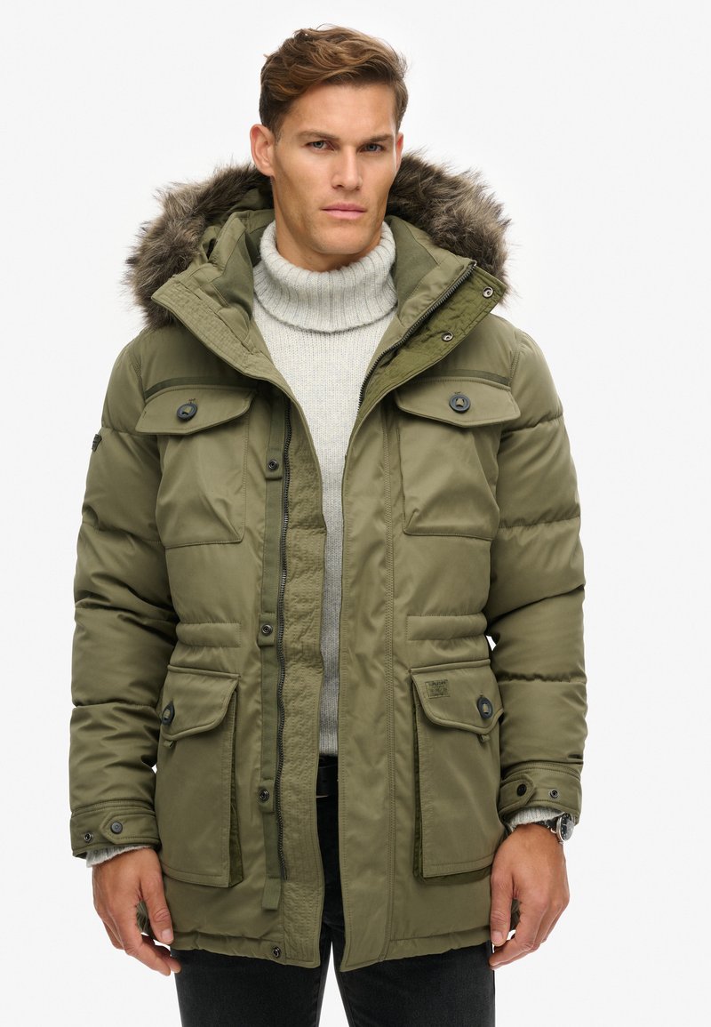 Chaqueta de plumas verde oliva con capucha forrada de piel, bolsillos abotonados y textura acolchada. Presenta un herraje oscuro y cierre con cremallera.