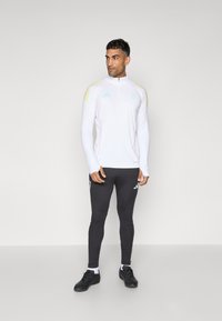 Witte lange mouwen sporttop met gele en roze accentkleuren, gecombineerd met zwarte leggings met een logo en strepen, gedragen met zwarte sportschoenen.