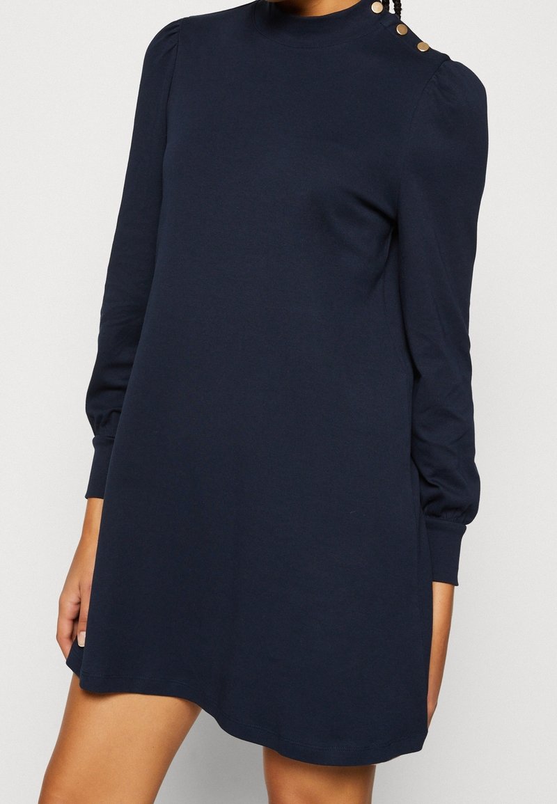 Femme portant une robe bleu marine à manches longues avec des épaules bouffantes et des boutons dorés sur l'épaule gauche.