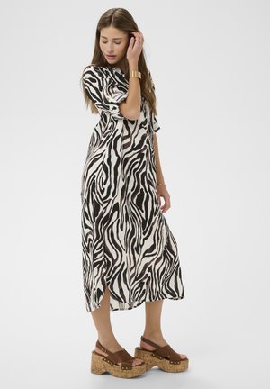 Naine seisab musta-valge sebratriibulise midi-kleit seljas, millel on käärvolditud varrukad, ja pruuni platvormsandaalidega, mille tallad on korki.