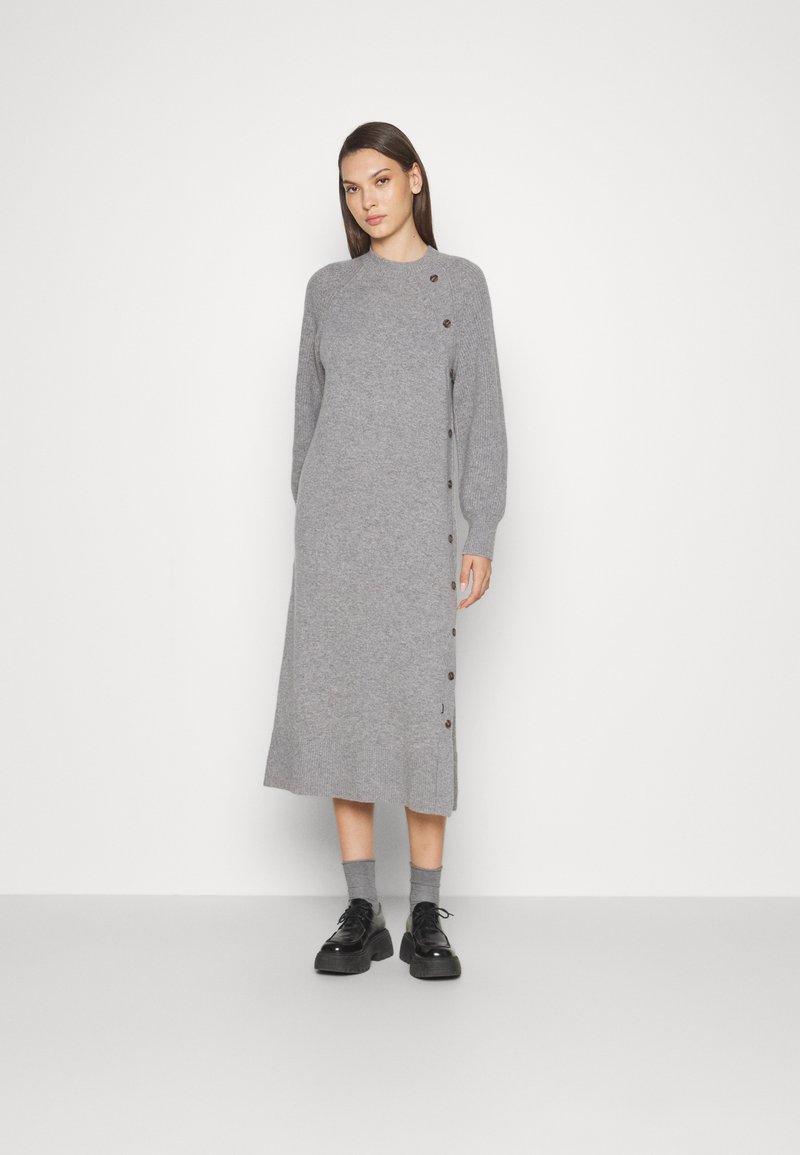 Szara dzianinowa sukienka z okrągłym dekoltem, długimi rękawami, detalem guzików po bokach i prostym krojem. Noszona z czarnymi butami i szarymi skarpetkami.