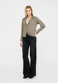 Olivgrüne Bluse mit langen Ärmeln und einem geknöpften Kragen; kombiniert mit hoch taillierten, weit geschnittenen schwarzen Jeans und schwarzen Stiefeln.