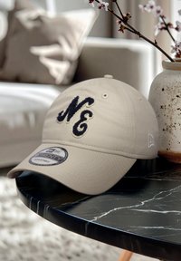 Căciulă din bumbac bej cu logo brodat "NE" în nuanță de bleumarin, cu vizieră curbată și ajustabilă, expusă pe o masă din marmură neagră.