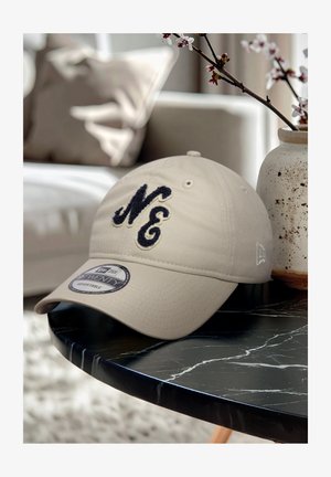 Beige Baumwollmütze mit einem navyblauen gestickten "NE"-Logo, gebogenem Schild und verstellbarem Sitz, auf einem schwarzen Marmortisch präsentiert.