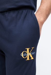 Høyre hånd i lommen på marineblå Calvin Klein joggebukser med gul og hvit CK-logo på låret, mot en ensfarget bakgrunn.