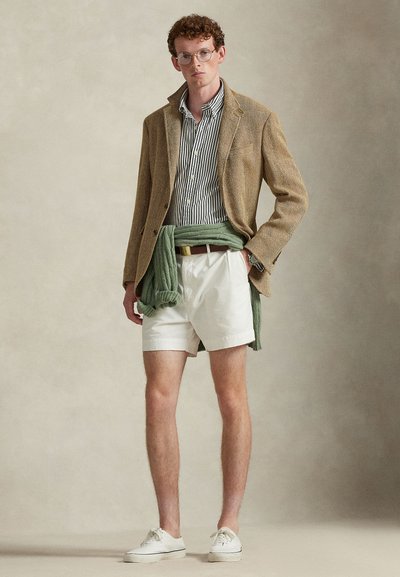 Μπλέιζερ από καφέ υφασμάτινη υφή, ριγέ πουκάμισο, λευκό σορτς και πράσινο πουλόβερ δεμένο στη μέση. Λευκά αθλητικά παπούτσια ολοκληρώνουν το σύνολο.