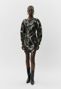 Moteris stovi vilkėdama juodą raštuotą ilgasleeve mini suknelę ir juodas smailias batus prieš paprastą šviesų foną.