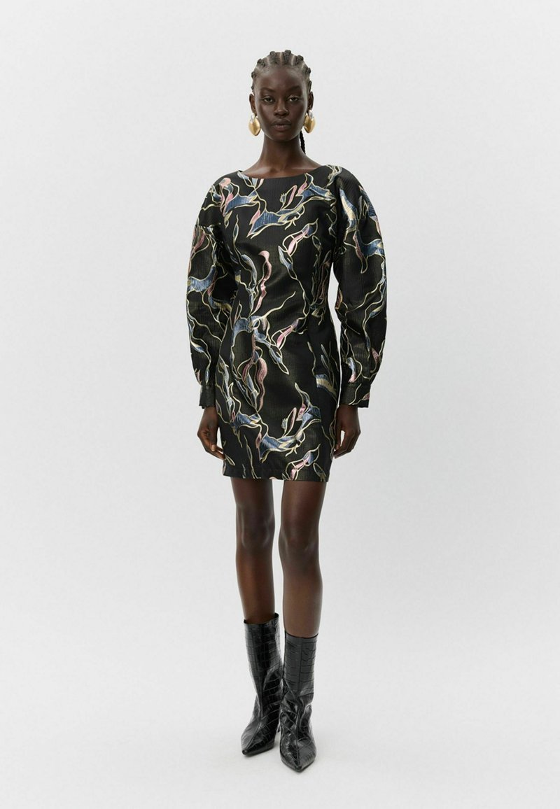 Moteris stovi vilkėdama juodą raštuotą ilgasleeve mini suknelę ir juodas smailias batus prieš paprastą šviesų foną.