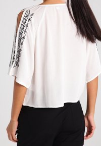 Hvid kortærmet bluse med sorte broderede detaljer langs skuldrene. Blødt, letstof med et løst, kort design.