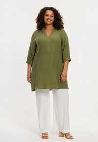 Olijfgroene, textuur tuniek met een V-hals en kraalaccenten, gecombineerd met witte, wijde broekspijpen en lichtbruine sandalen met hakken.