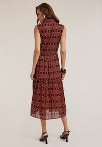 Red patterned dress with a high collar, sleeveless design, elastic waist, and tiered skirt. Accented with chunky bracelets and heeled sandals.