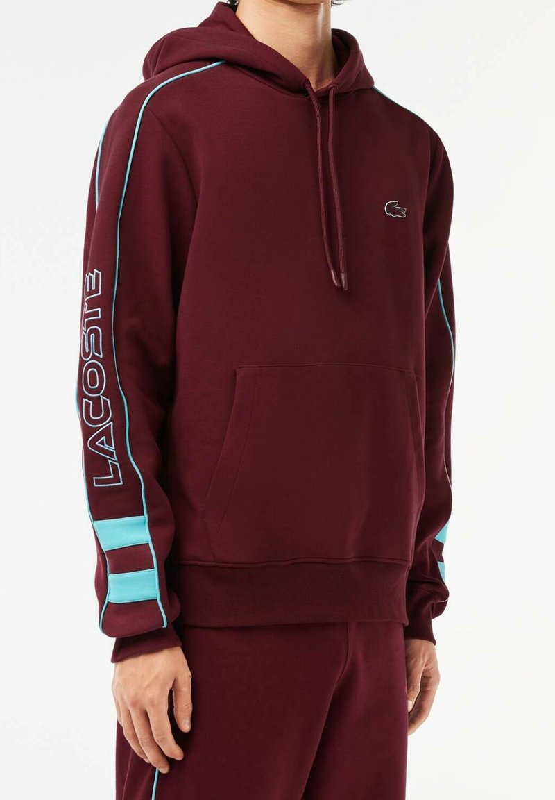 Sweat à capuche bordeaux avec une poche kangourou, présentant des accents bleu clair et le logo 'LACOSTE' sur la manche, confectionné en matériau doux.