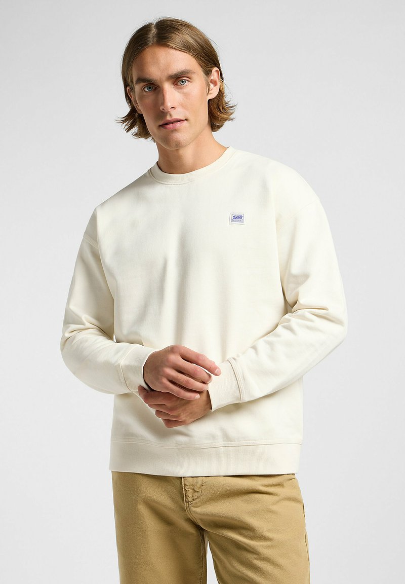 Sweatshirt crème en tissu doux, avec un col rond, des poignets côtelés et un petit patch logo bleu et blanc sur la poitrine.