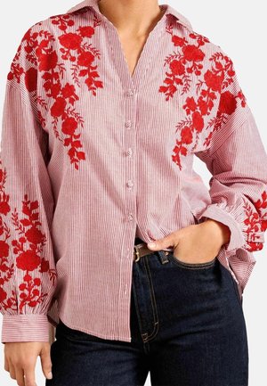 Chemise à manches longues à boutons avec broderie florale rouge sur tissu rayé rose, associée à un jean bleu foncé et une ceinture marron.