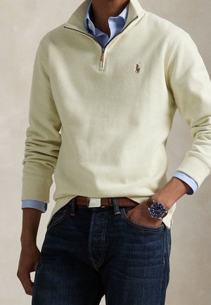 Man wearing a beige quarter-zip sweater over a blue button-up shirt, dark jeans, brown belt, and a blue wristwatch with a metal band.