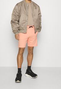 Veste bomber taupe légère sur une chemise beige, assortie à un short rose vif et des baskets noires avec des semelles blanches.