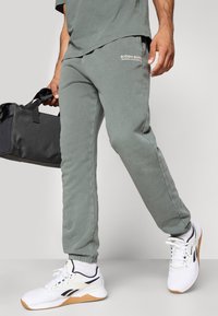 Πράσινο βαμβακερό παντελόνι φόρμας με ελαστικούς μανσέτες, με λευκό λογότυπο στο μπροστινό μέρος. Συνδυασμένο με λευκά αθλητικά παπούτσια και μαύρη τσάντα duffel.