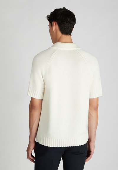 Uomo che indossa un maglione a maniche corte di cotone off-white con colletto a coste, polsini e orlo, in piedi contro uno sfondo chiaro e semplice.