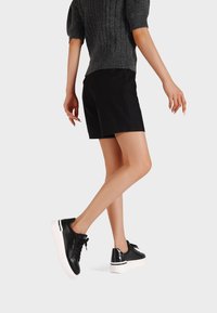 Persona che indossa pantaloni corti neri, un maglione lavorato a maglia grigio scuro e scarpe da ginnastica nere basse con suole bianche, che cammina su uno sfondo uniforme.