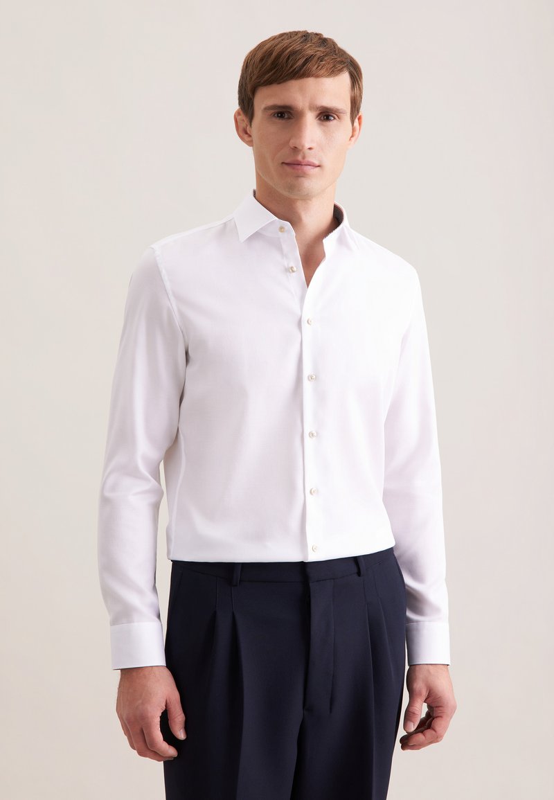 Chemise blanche à manches longues avec col classique, texture douce et boutons beiges, associée à un pantalon foncé plissé.
