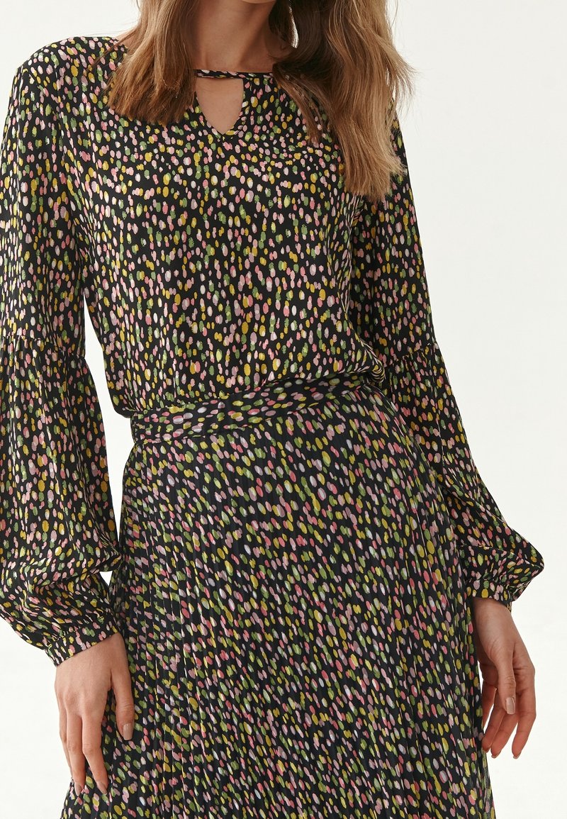 El conjunto de blusa y falda negra presenta un patrón salpicado de colores en rosa, amarillo y verde, con mangas largas y un detalle de escote en forma de ojo de cerradura.