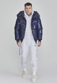 Navy puffer jacket with shiny, quilted texture and hood, paired with a light gray sweatshirt and matching sweatpants, and beige sneakers.