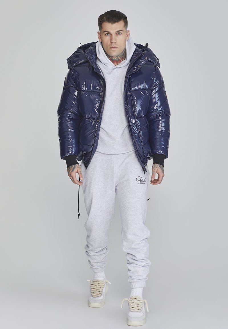 Navy puffer jacket with shiny, quilted texture and hood, paired with a light gray sweatshirt and matching sweatpants, and beige sneakers.