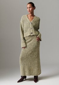 Zeleni pletivi džemper s V-izrezom i tri velika gumba, uparen s odgovarajućom dugom suknjom. Teksturirani materijal i prilagođene rukave. Smeđe gležnjače.