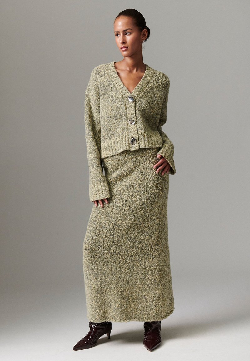 Zeleni pletivi džemper s V-izrezom i tri velika gumba, uparen s odgovarajućom dugom suknjom. Teksturirani materijal i prilagođene rukave. Smeđe gležnjače.