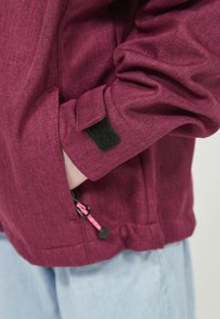 Main reposant dans la poche avant d'une veste rose foncé avec une tirette de fermeture éclair noire et rose et une bande velcro noire sur le poignet de la manche.