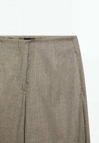 Pantalon beige taille haute avec fermeture éclair avant dissimulée et poches latérales, en tissu tissé texturé.