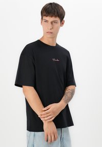 Young man with short brown hair wearing a black oversized T-shirt with small pink logo, light blue jeans, and tattoo on left forearm.