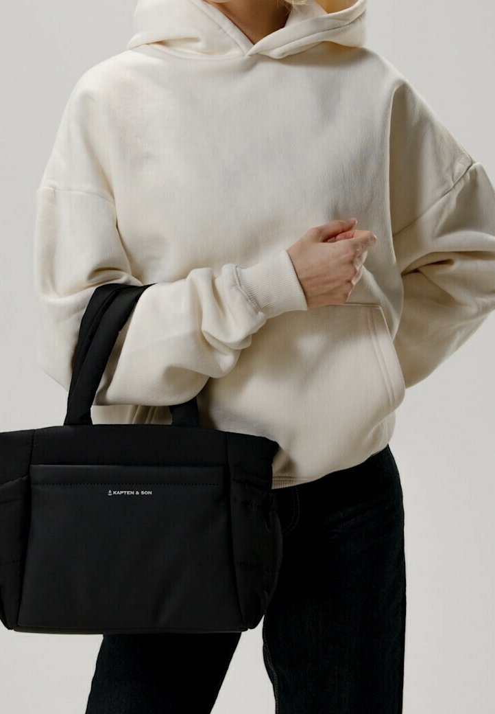 Svart toteväska med dubbla handtag, vadderad textur, framficka och diskret märkning, kombinerad med en vit oversized hoodie.