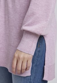 Pull en rose clair avec des poignets et un ourlet côtelés, doté de fentes sur les côtés. Associé à un jean en denim bleu, montrant un gros plan de la texture du tissu.