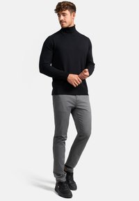 Pull à col roulé noir avec un design ajusté, associé à un pantalon gris à motifs et des baskets noires, pose debout avec les mains détendues.