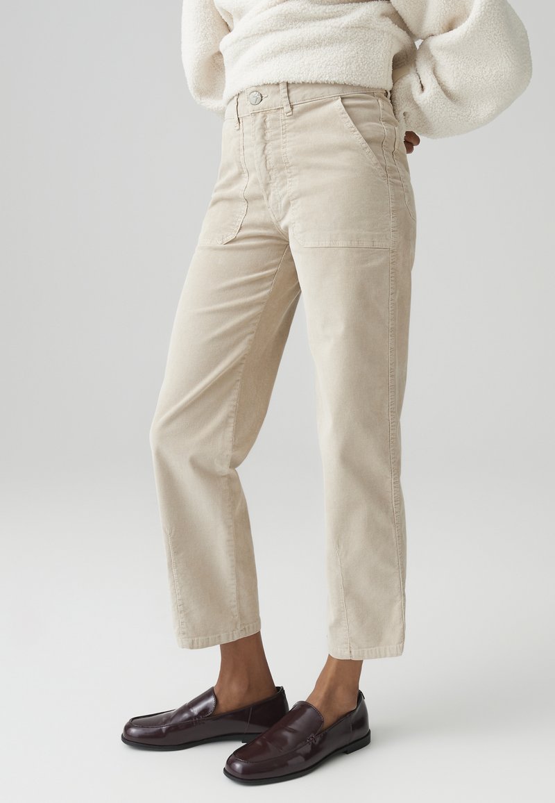 Pantaloni in corduroy beige con vita alta, tasche a toppa frontali e gamba dritta, abbinati a scarpe slip-on di un borgogna scuro.