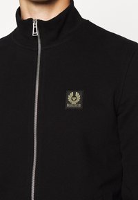 Svart zip-up jacka med hög krage, tillverkad av mjuk tyg, med en fyrkantig Belstaff-logga i guld på vänster bröstområde.