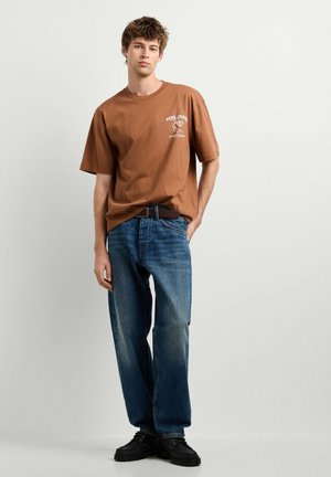 Giovane uomo che indossa una t-shirt marrone Pepe Jeans, jeans blu e scarpe nere, in piedi davanti a uno sfondo chiaro e uniforme.