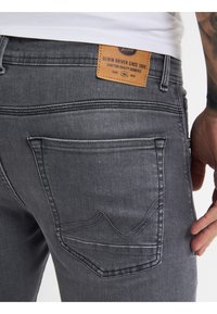 Vaqueros de mezclilla gris oscuro con un ligero desgastado, que presentan un diseño de bolsillo cosido y una etiqueta de cuero en la cintura con los detalles de la marca.
