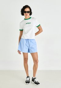 T-shirt bianco a maniche corte con dettagli verdi e testo "drive", abbinato a pantaloni corti azzurri chiari e sneakers nere con lacci bianchi.