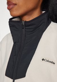 Polarowa kurtka z wysokim kołnierzem, z czarnym przednim panelem i kremowym ciałem, z zapięciem na zamek błyskawiczny oraz logo Columbia na boku.